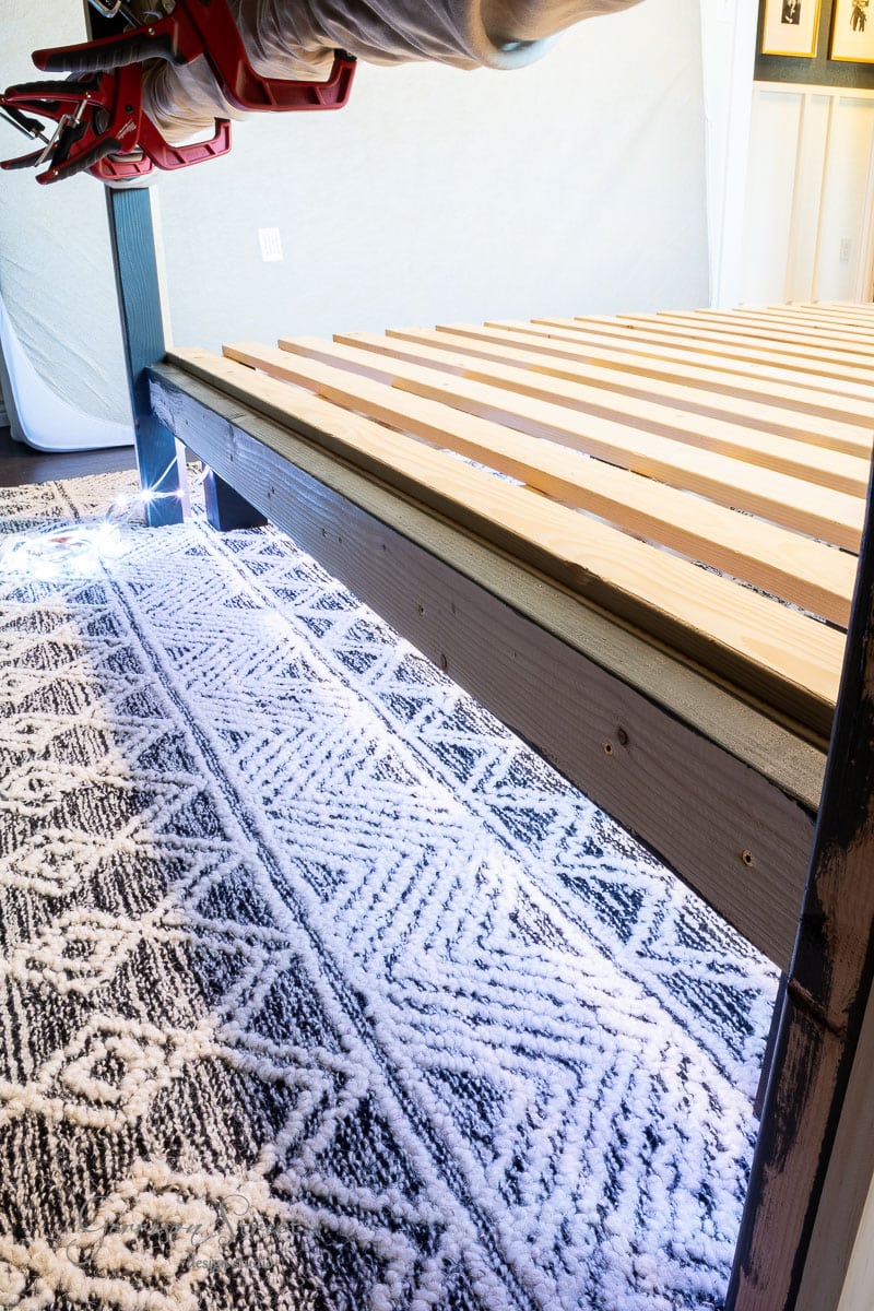 A modern DIY full size bed frame with wooden slats is illuminated by LED strip lights underneath. The carpet features an intricate geometric pattern in shades of gray and white, enhancing the stylish ambiance of the room.