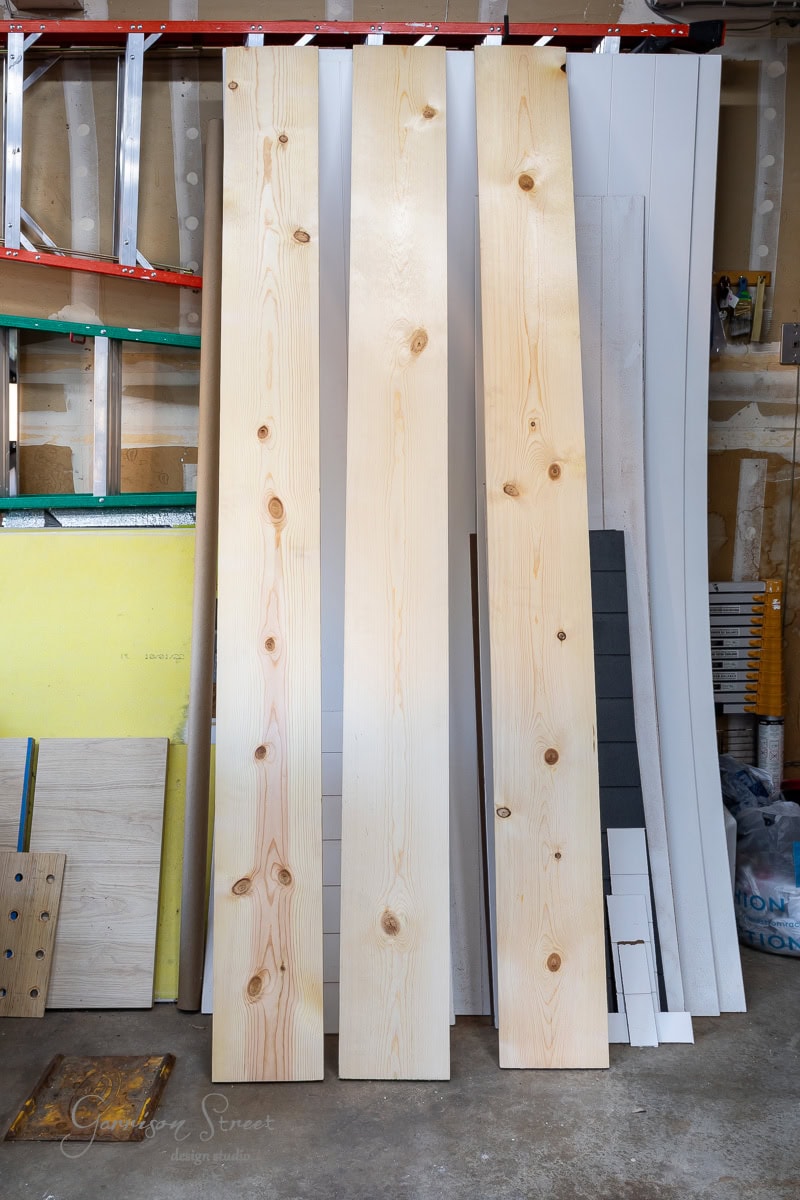 Three large, vertical wooden planks stand against a wall in a workshop, primed for a DIY full-size bed frame project. Surrounding them are various boards, a colorful ladder, and assorted building materials. The space has a utilitarian feel with items organized for easy access.