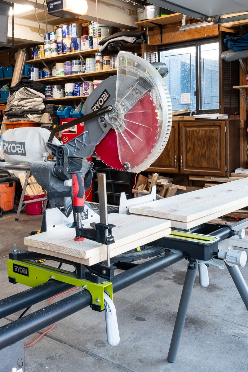 A miter saw with a red blade sits on a workbench in a cluttered garage, ready for action. Wooden planks await in front—perfect for crafting that DIY full-size bed frame. Shelves brimming with paint cans and tools paint the background with possibilities.