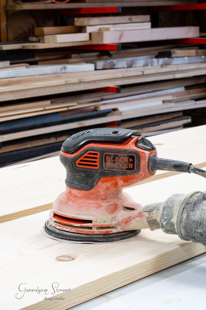 A black and orange Black & Decker orbital sander sits on a wooden plank, surrounded by stacks of lumber in a workshop, ready to smooth the edges for your next DIY full-size bed frame. The sander is slightly dusty from use.