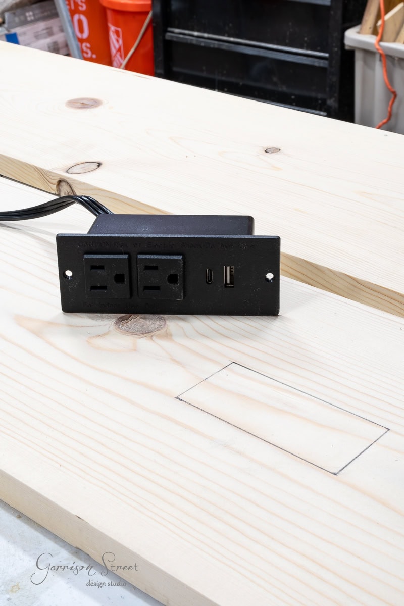 A black power outlet and USB port panel rests on a wooden table, amidst tools hinting at a DIY full-size bed frame project. The panel has one closed and one open socket, along with two USB ports. A pencil outline marks the wood, suggesting an imminent installation spot.