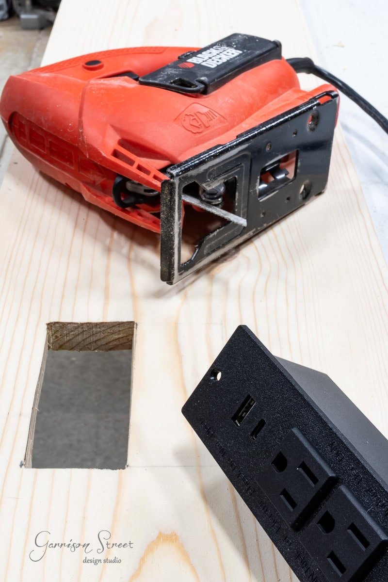 A red jigsaw rests on a wooden board with a rectangular cutout, ready for the next DIY project. Nearby, a black electrical outlet fixture awaits installation. The board, marked with the "Garrison Street Design Studio" logo, hints at its potential role in crafting a full size bed frame masterpiece.