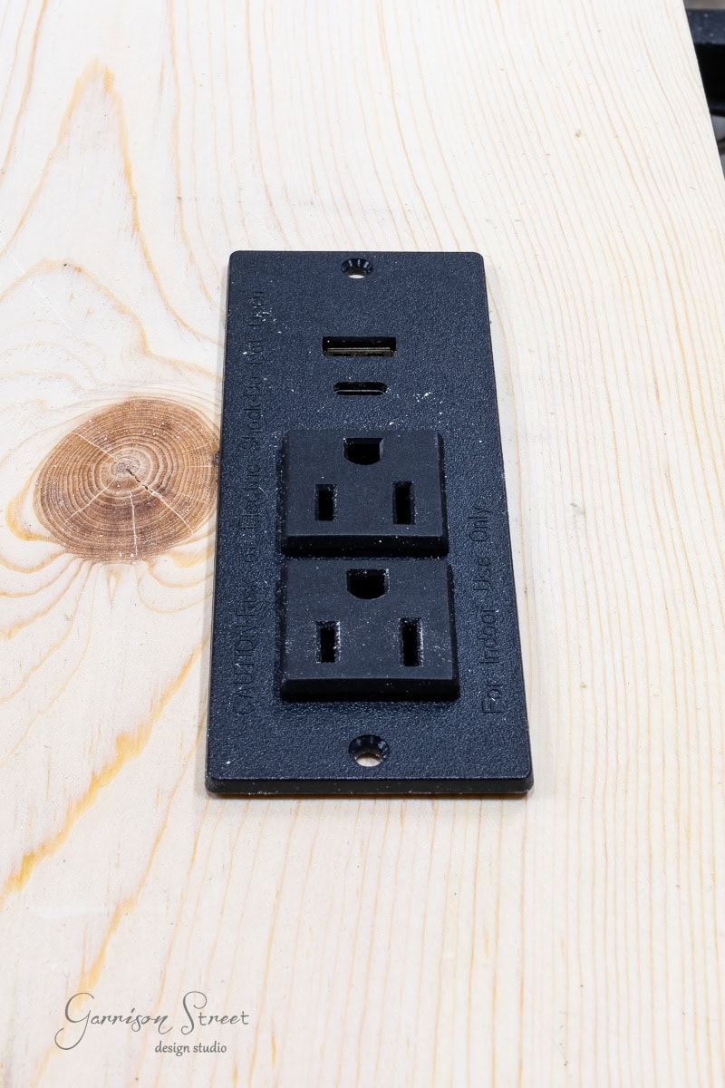 A black electrical outlet with two standard sockets and two USB ports is seamlessly integrated into a light wood bed frame. The wood showcases a beautiful knot pattern and grain texture, perfect for those who enjoy DIY projects to customize their full-size bed frame experience.