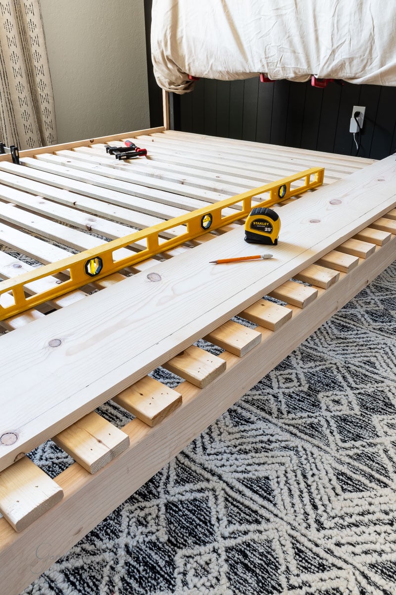 A DIY full-size wooden bed frame is in the process of being assembled, with slats and tools like a yellow level, tape measure, and pencil resting on it. The bed sits in a room adorned with a patterned rug on the floor.