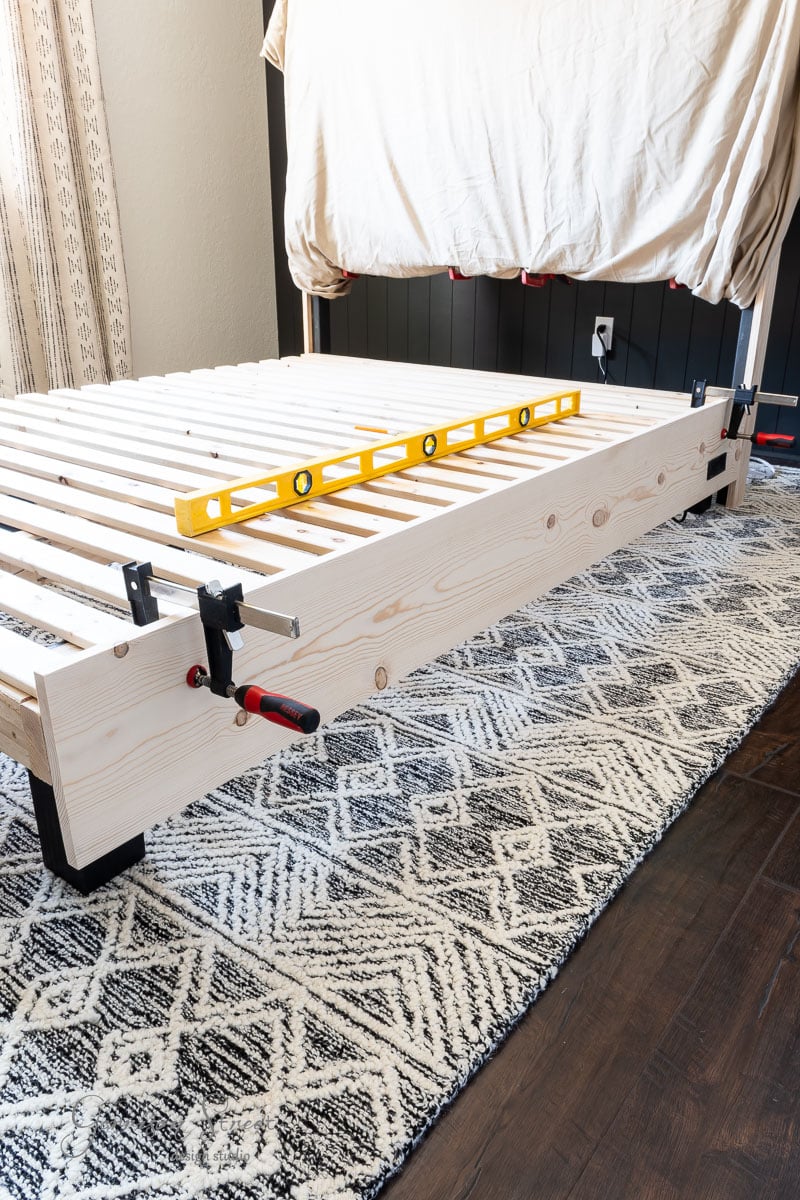 A DIY full-size bed frame is under construction, secured with clamps. A yellow level ensures precise alignment. The setup rests on a patterned area rug, with a curtain providing the perfect backdrop.
