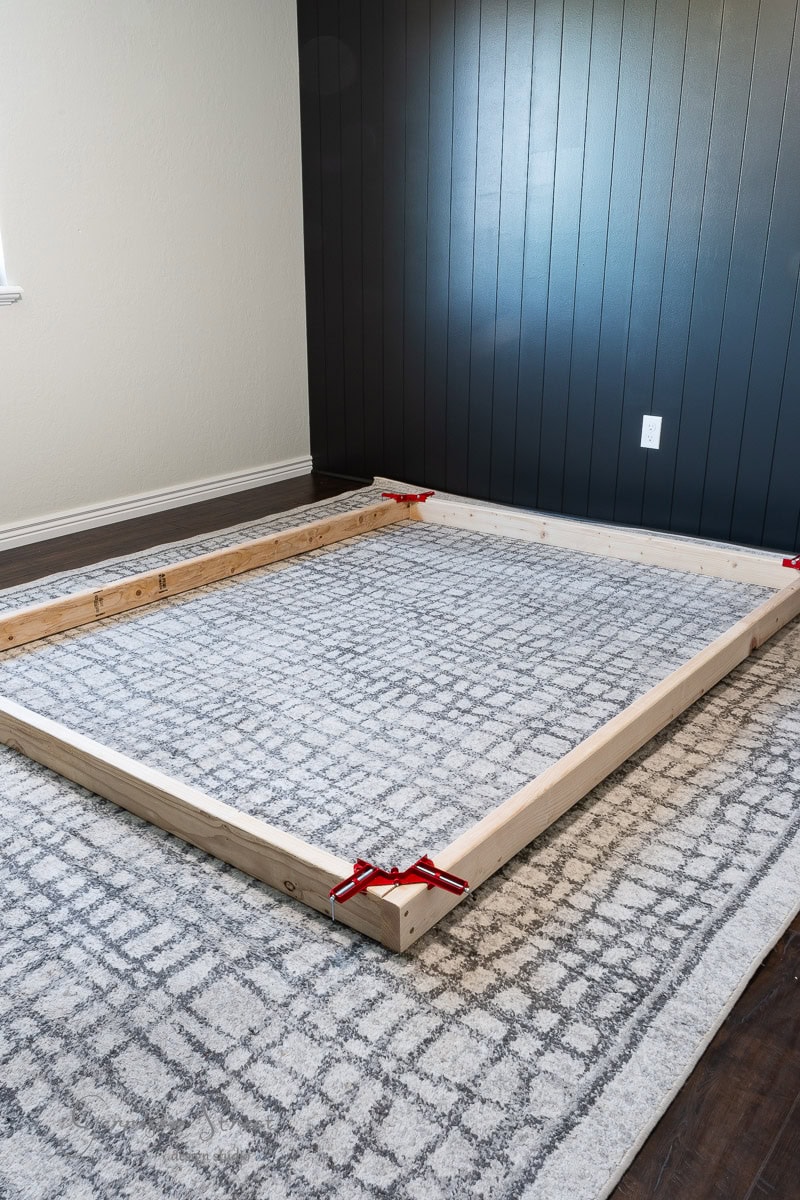 A wooden frame for a DIY full-size bed is held together with red corner clamps, resting on a large patterned rug in a room with dark wall paneling.
