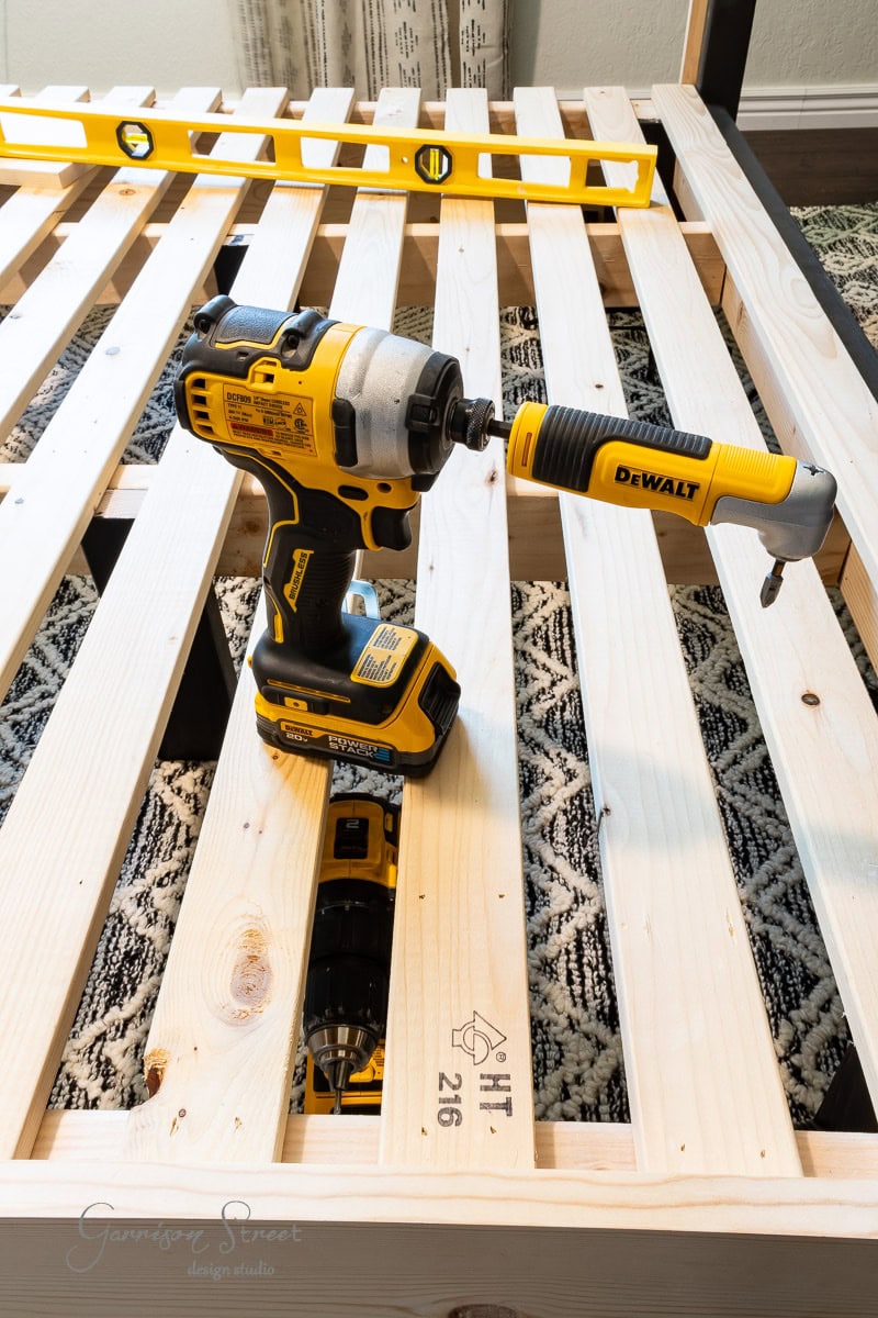A DeWalt power drill is placed on a wooden full size bed frame with slats. A yellow level rests on the frame, capturing the essence of a DIY project indoors.