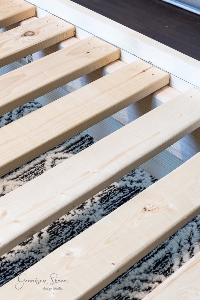 Close-up of a wooden bed frame, beautifully crafted as a DIY full-size project, with unfinished wooden slats resting on a patterned rug. The logo "Garrison Street Design Studio" is visible in the bottom left corner.
