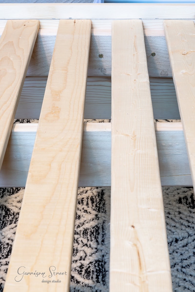 Close-up of a DIY full-size wooden bed frame with slats laid horizontally. The wood has a light, natural finish, and the bed rests gracefully over a patterned black and white carpet.