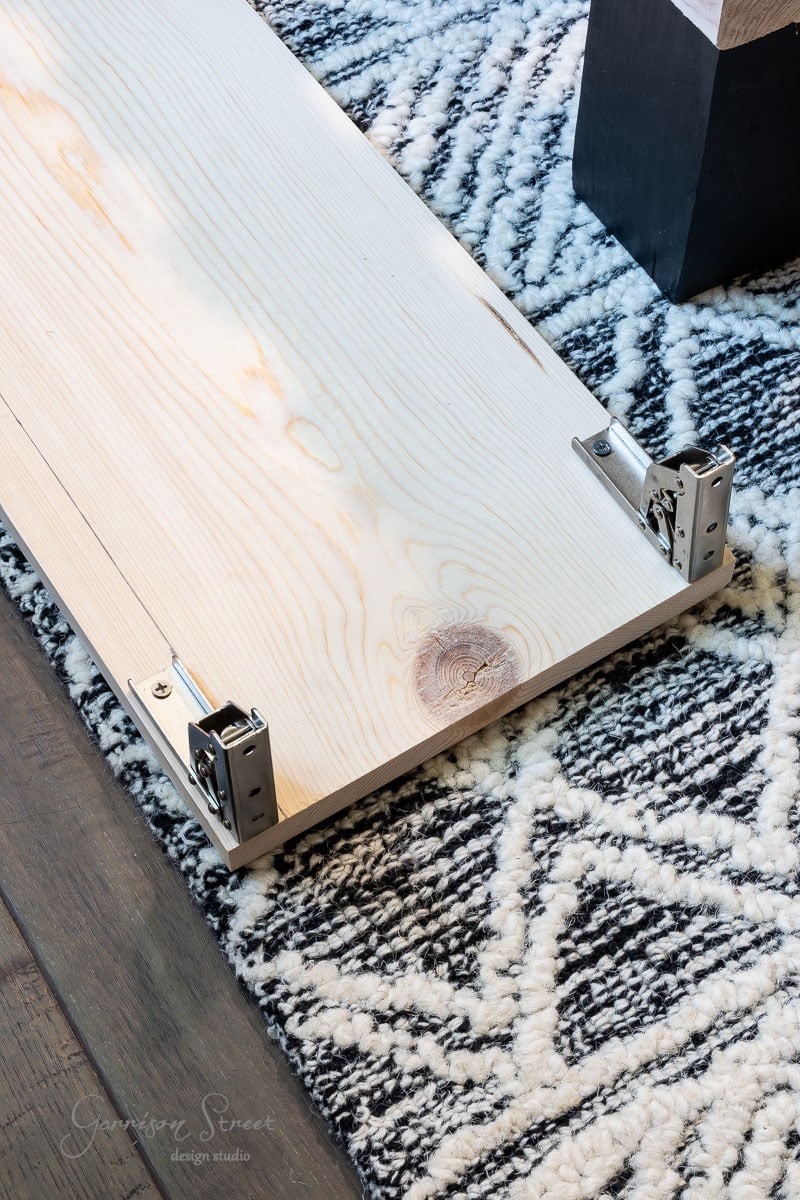 A wooden plank with metal brackets rests on a patterned rug, hinting at a DIY full-size bed frame in progress. The brackets secure one end of the plank, fitting perfectly with furniture assembly plans. The rug showcases a striking geometric black and white design.