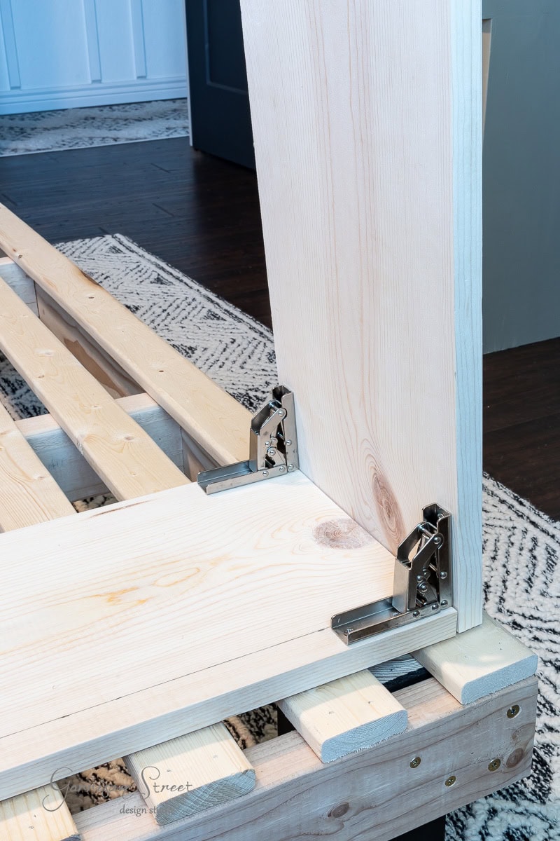 Close-up of DIY full-size wooden bed frame assembly, showing metal brackets securing a vertical and horizontal plank. The frame rests on a patterned rug, with a partially visible wall in the background.