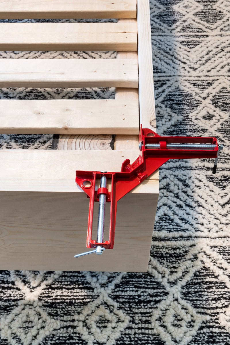 A red woodworking corner clamp secures two pieces of light-colored wood on a geometric-patterned carpet. The wood looks like part of a DIY full-size bed frame project, with visible slats on top, suggesting a sturdy and stylish design in progress.