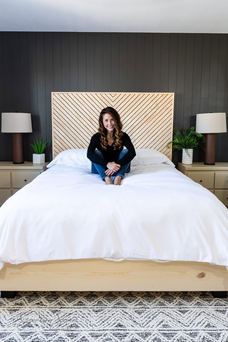 A person sits cross-legged on a bed with a white duvet, nestled within a stylish DIY full-size bed frame. The room features a dark accent wall and wooden headboard with a chevron pattern. Two lamps and small plants adorn the bedside tables, while a patterned rug covers the floor.