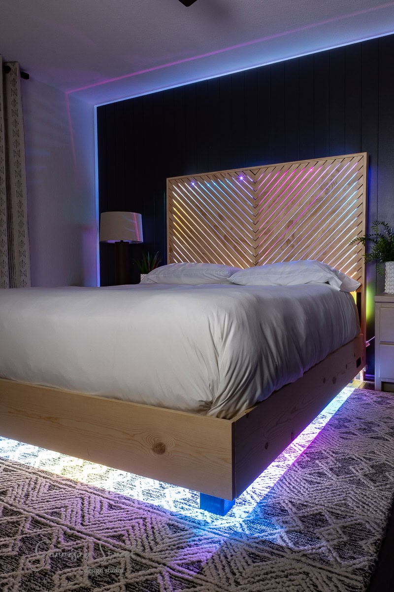 A modern bedroom showcases a DIY full-size bed frame with a wooden structure and headboard. Colorful LED lights illuminate the base, accentuating the headboard. The room is adorned with a patterned rug, bedside table, lamp, and a plant for added charm.