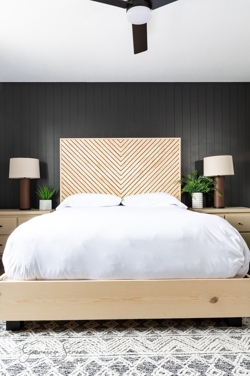 A bedroom with a modern design showcases a DIY full-size bed frame with a white comforter. The headboard flaunts a chevron pattern, while two nightstands with lamps flank the bed. A patterned rug graces the floor, and a dark accent wall adds striking contrast.