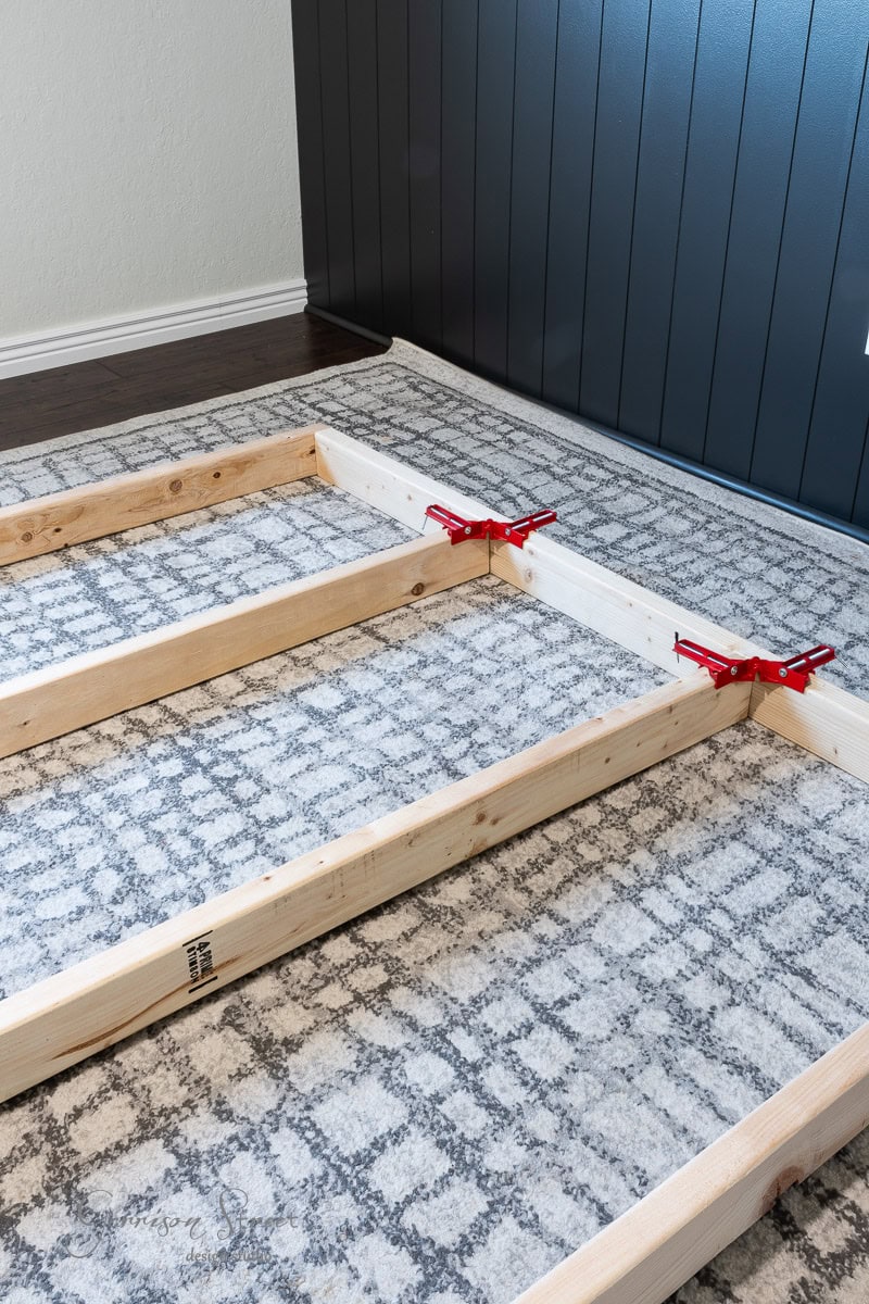 Wooden frames with red clamps are assembled on a carpeted floor, adjacent to a navy blue paneled wall, as part of a DIY full-size bed frame project.