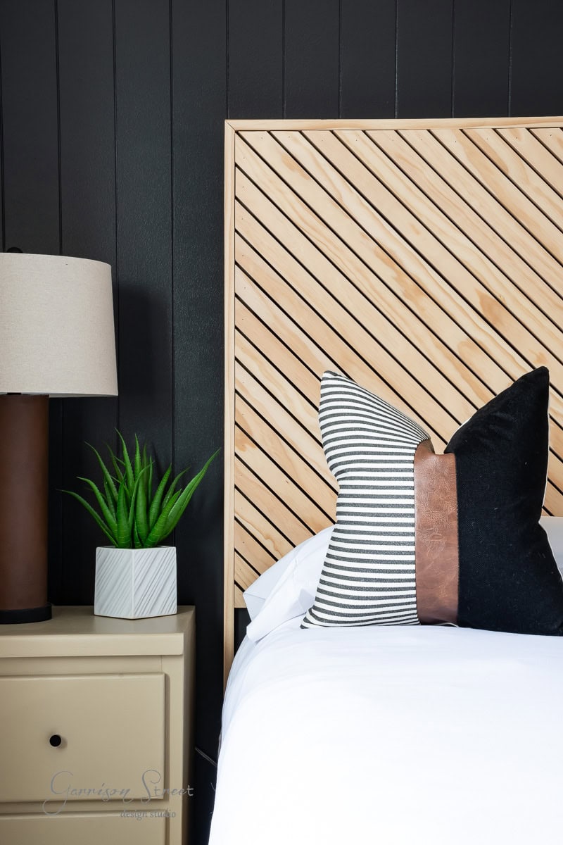 Modern bedroom with a diagonal wooden slat headboard on a DIY full-size bed frame, black accent wall, and a neatly made bed. A beige nightstand holds a potted plant and a lamp with a beige shade. Pillows on the bed feature black, white, and leather textures.