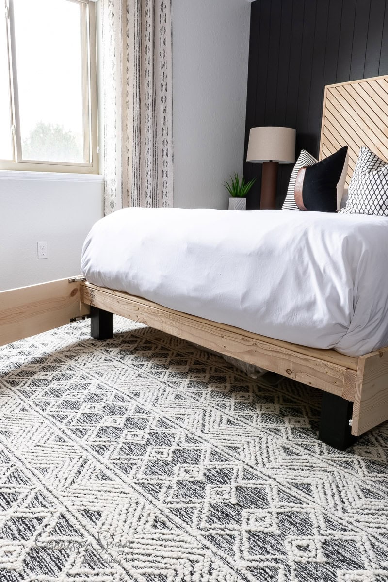 A modern bedroom showcases a DIY full-size bed frame with a patterned headboard, draped in crisp white linens. A geometric rug enhances the floor, while a window with curtains and a small plant on the nightstand complete the serene space.