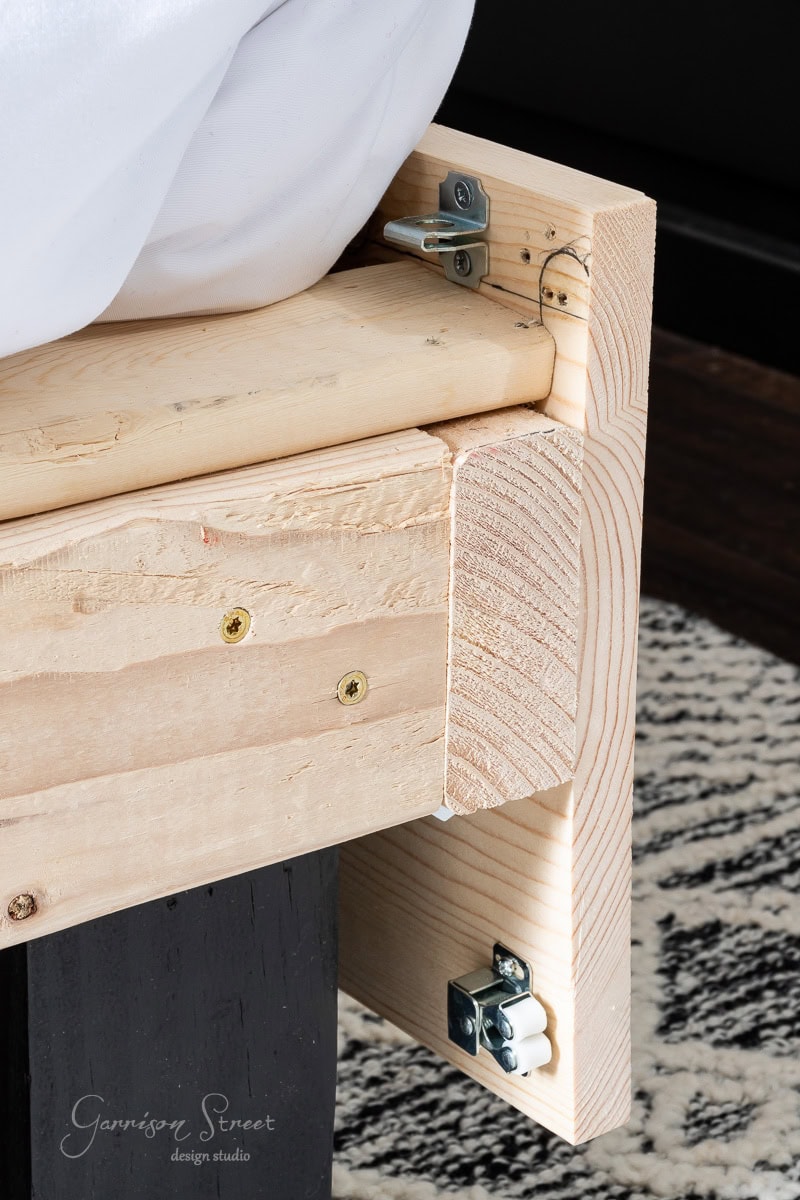 Close-up of a corner of a DIY full-size bed frame on a textured rug. The frame displays unfinished wood with visible screws and corner brackets, showcasing detailed joinery. A portion of white fabric, likely a bedsheet, is seen on top.