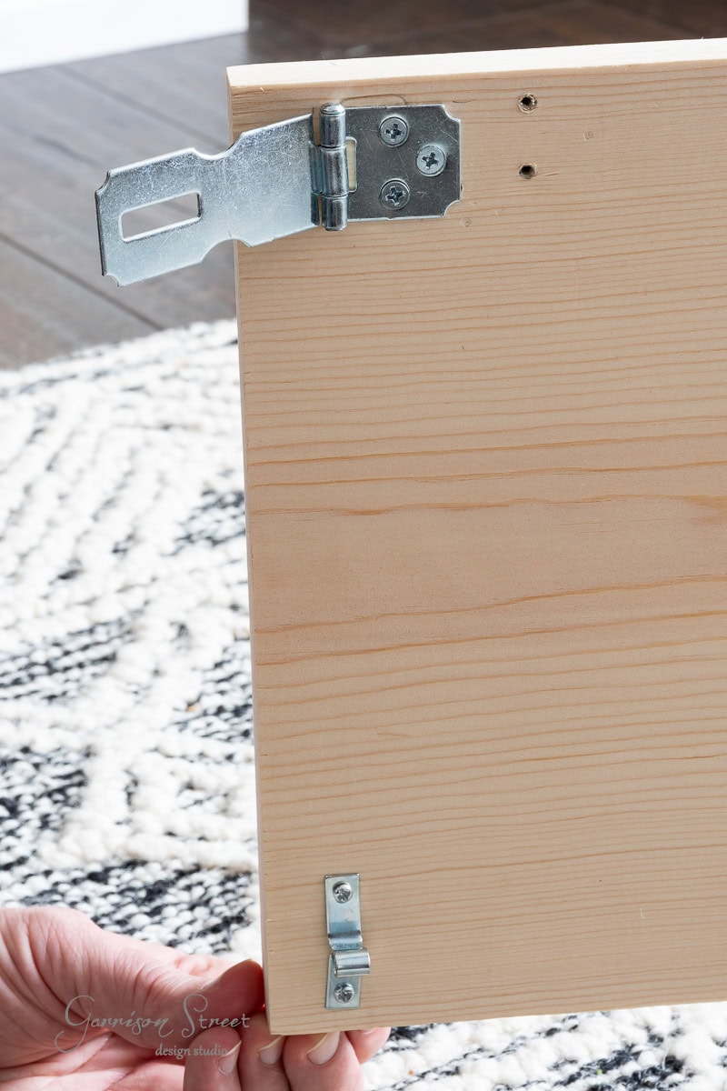 A hand holds a wooden board with metal hinges attached, likely the start of a DIY full-size bed frame. The board rests on a patterned carpeted floor, hinting at the creativity and craftsmanship involved in building functional, custom pieces for your home.