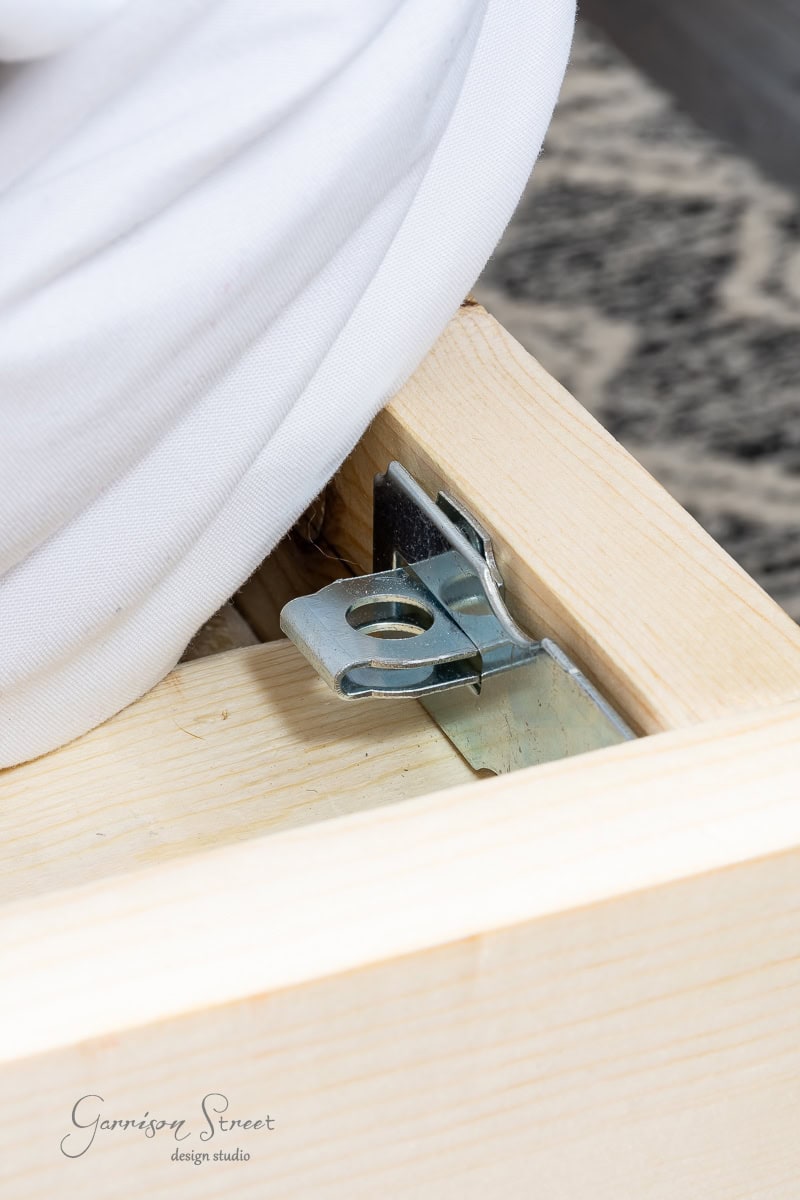 Close-up of a DIY full-size wooden bed frame corner, showcasing a metal bracket and bolt partially visible beneath a white mattress. The textured gray and white carpet on the floor adds a cozy touch.