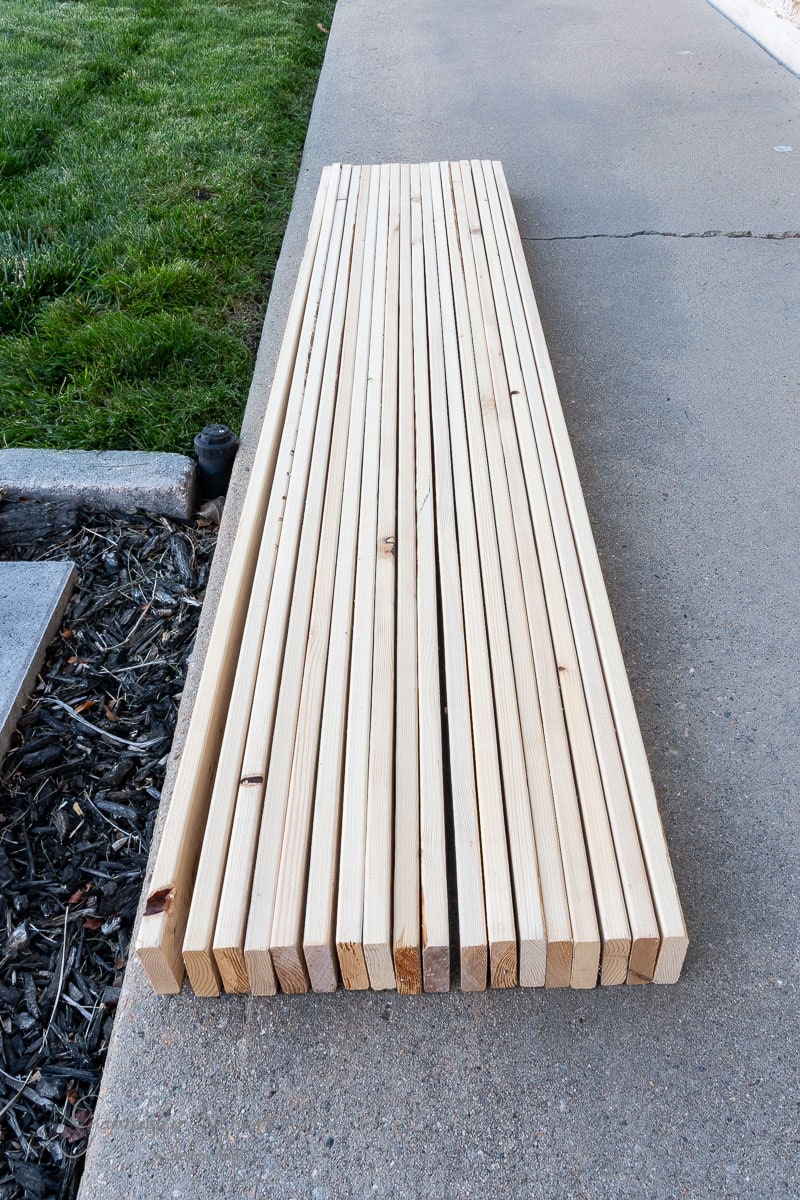 A stack of wooden planks, perfect for a DIY full-size bed frame, is neatly arranged on a concrete surface next to a grassy area. The planks are uniform in size and color, with natural wood grain visible.