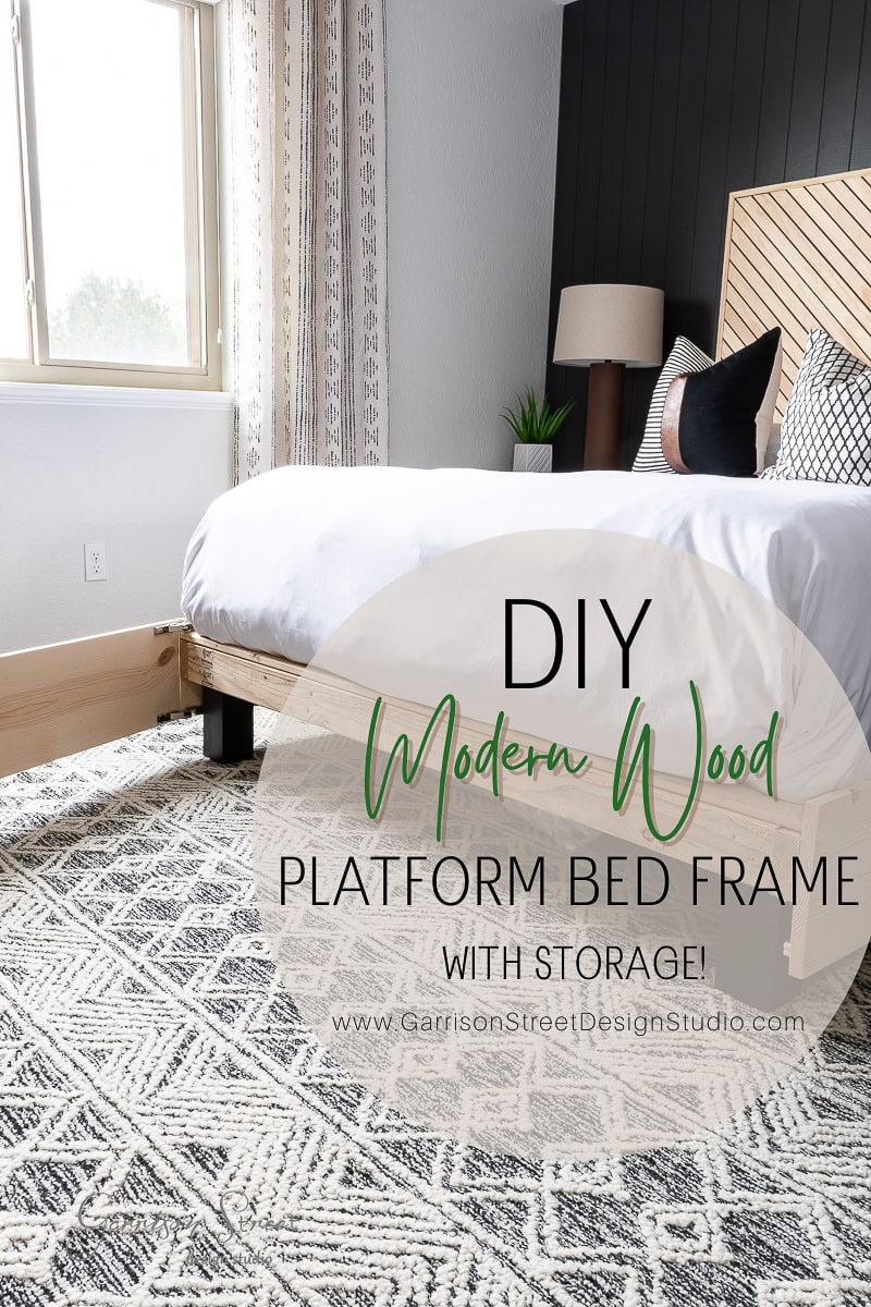 A modern bedroom showcases a DIY full size platform bed frame made of light wood, complete with storage underneath. The bed is dressed in white and gray linens, while a geometric patterned rug adorns the floor. Sunlight streams through a large window, highlighting this stylish and functional design.