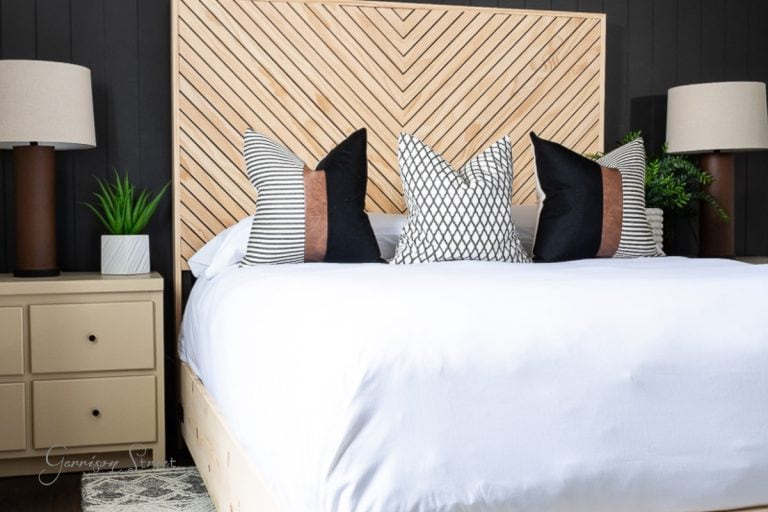 A modern bedroom features a wooden headboard with a chevron pattern, part of a DIY full-size bed frame. The neatly made bed showcases a white cover and four stylish pillows in black, white, and brown hues. Two lamps and a small plant rest on the bedside tables.