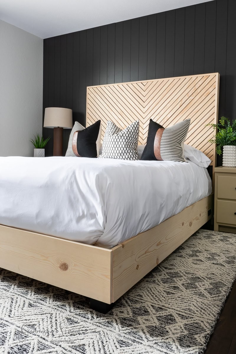 A modern bedroom featuring a DIY wooden queen bed with a herringbone-patterned headboard. The bed is made with white linens and decorative pillows. A black accent wall complements a lamp and potted plants on side tables, with a patterned rug on the floor.