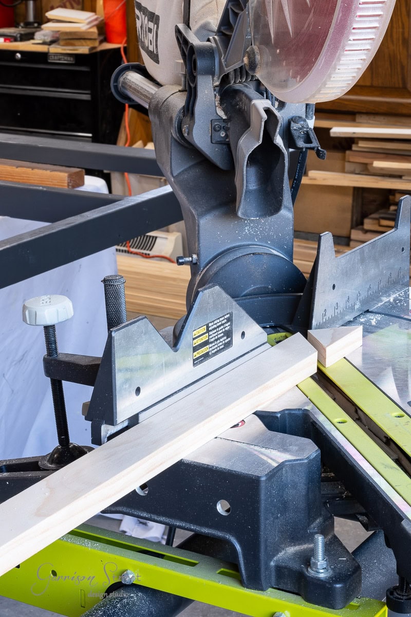 A miter saw positioned in a workshop, with a wooden plank on the saw table, hints at the start of a DIY headboard project. Surrounded by tools and wood pieces, the scene captures the essence of woodworking or carpentry.