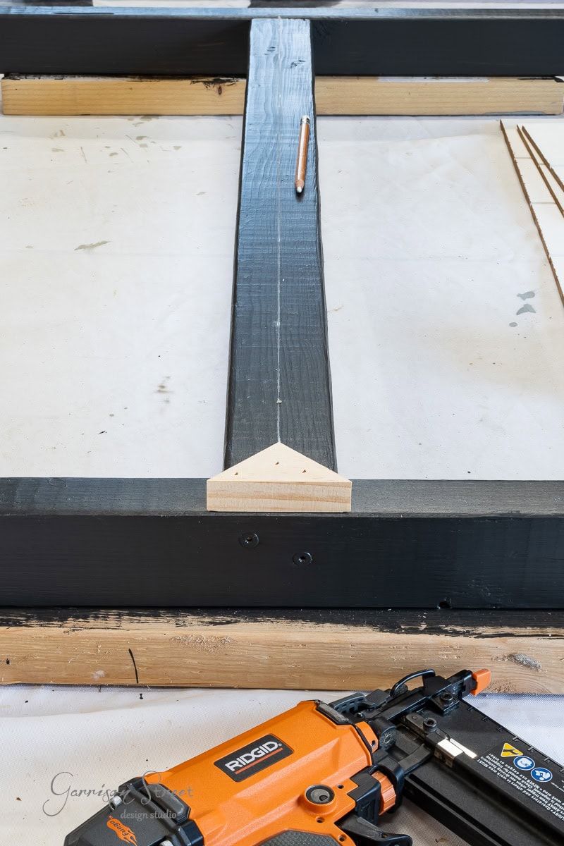 A carpentry workbench with a large black wooden frame and an attached wooden triangular block. Near the frame rests a pencil, while an orange nail gun lies nearby—perfect for your next DIY project, whether crafting a headboard or any queen-sized masterpiece.