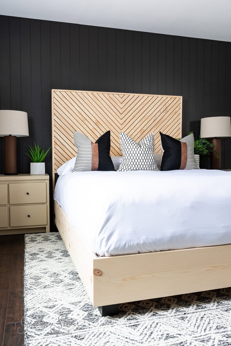 A modern bedroom showcases a DIY wooden herringbone-patterned headboard for a queen bed. Black and white pillows embellish the bed, flanked by nightstands with matching lamps and green plants. Dark walls complement the stylish ensemble, while a patterned rug completes the aesthetic.