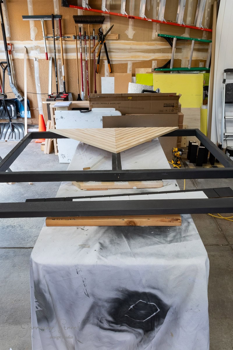 A workshop with a wooden headboard project in progress on a table. A black frame and a triangular wooden design are visible. Various tools and materials, including ladders and boxes, enhance the DIY atmosphere in the background.