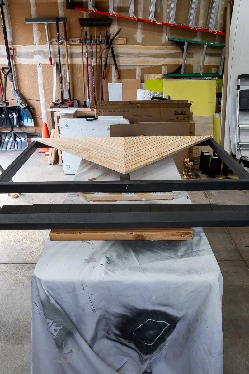 A garage workspace with various tools and equipment. In the foreground, there's an unfinished DIY wooden headboard for a queen bed on a table covered with a white cloth. Ladders and garden tools hang on the wall in the background.