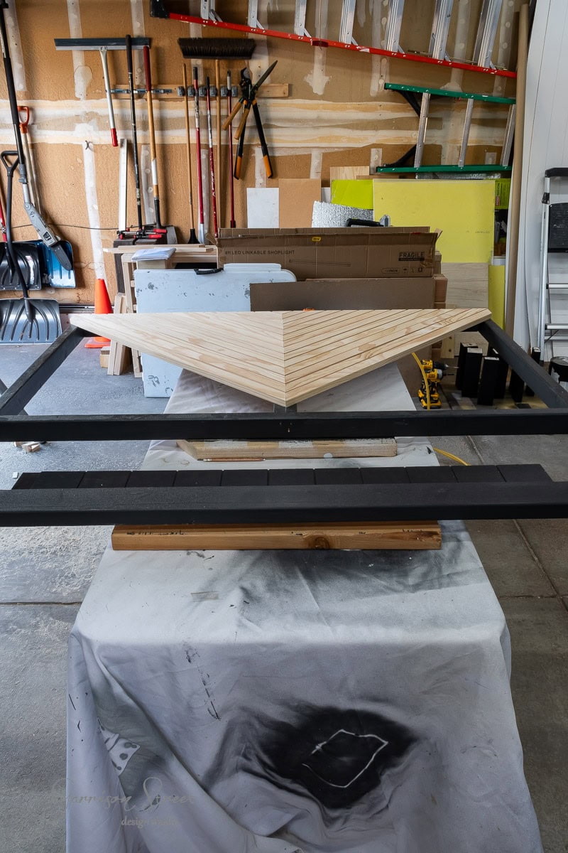 A garage workspace with shelves of tools and equipment showcases DIY creativity. In the foreground, a triangular wooden piece with a chevron pattern hints at becoming a unique headboard. Resting on a black metal frame atop a draped table, various boards and materials are stacked against the wall.
