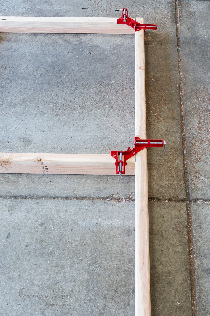 Wooden beams are clamped together at a right angle with red corner clamps, forming a frame perfect for a DIY headboard. The setup rests on a concrete floor, with sawdust scattered around the edges.