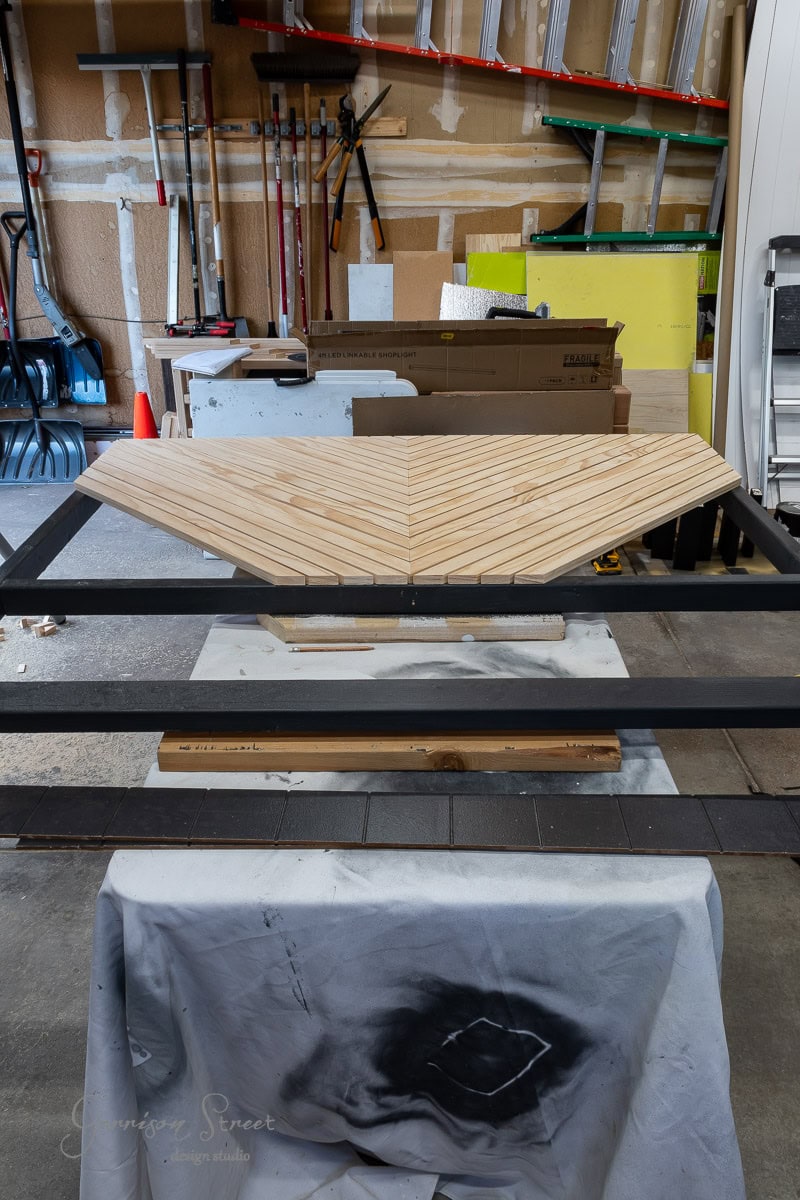 In a garage workshop, a DIY woodworking project is underway. A partially assembled headboard with a herringbone pattern rests on a large black frame, perfect for a queen-sized bed. Tools and materials are neatly scattered in the background, setting the stage for creativity.
