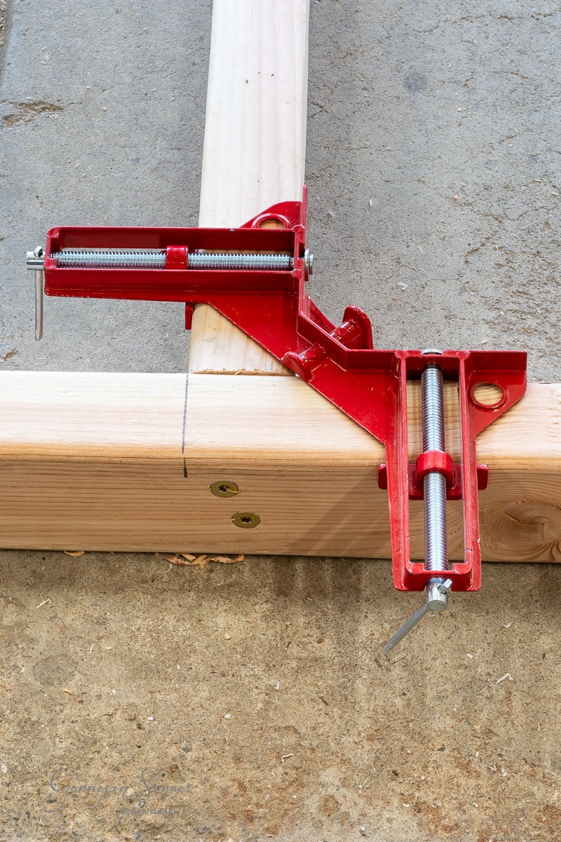 A red corner clamp secures two wooden pieces in an L-shape on a workshop floor, ideal for a DIY project like crafting a headboard for a queen-sized bed. The clamp's metal screws are visible against the textured concrete surface, ensuring precision and stability.