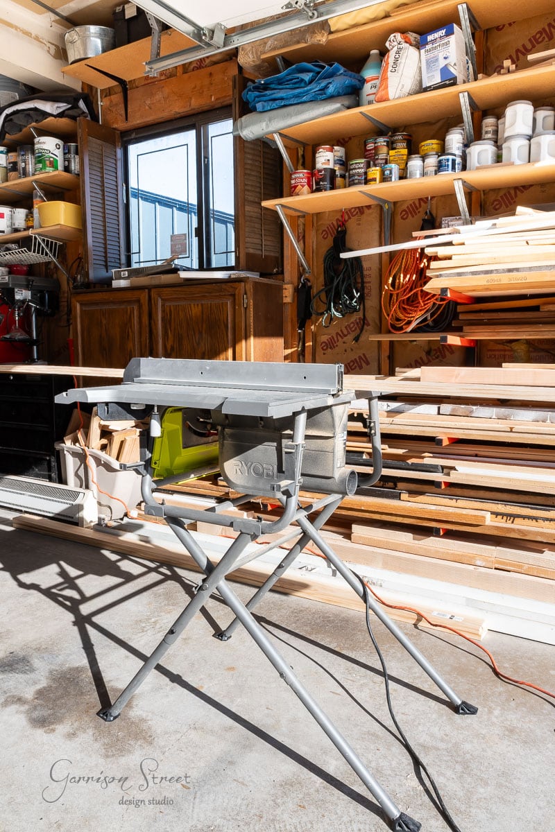 A workshop with a portable Ryobi table saw at its heart, framed by shelves holding tools, paint cans, and materials. Sunlight streams through a window, illuminating the organized space as shadows dance across the concrete floor. Plans for a DIY queen headboard are sketched out nearby.