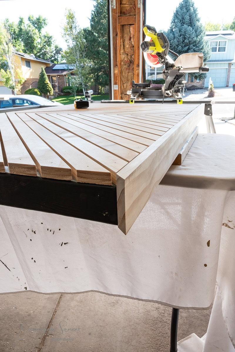 A woodworking project unfolds in a garage, featuring a partially assembled queen headboard with slats. A miter saw sits ready on the workbench nearby. Sunlight illuminates the scene, revealing trees and houses beyond, perfectly setting the stage for this DIY endeavor.