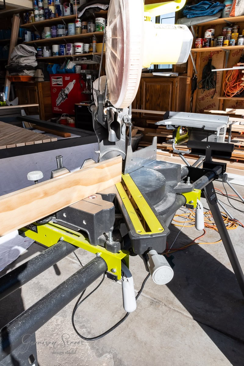 A compound miter saw in a workshop is positioned to cut a wooden board, perfect for your queen DIY headboard project. The bright light highlights the blade and the wood, while shelves stocked with various tools and supplies fill the background.