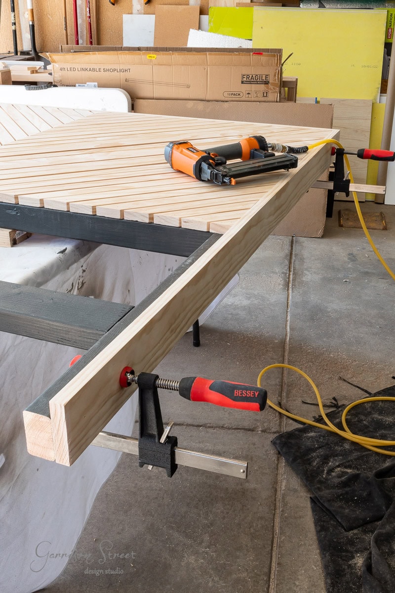 A wood workshop scene displays a large wooden panel, possibly a DIY queen headboard, clamped to the workbench. Two pneumatic nail guns rest atop the panel with hoses attached, while various tools and materials are scattered in the background.