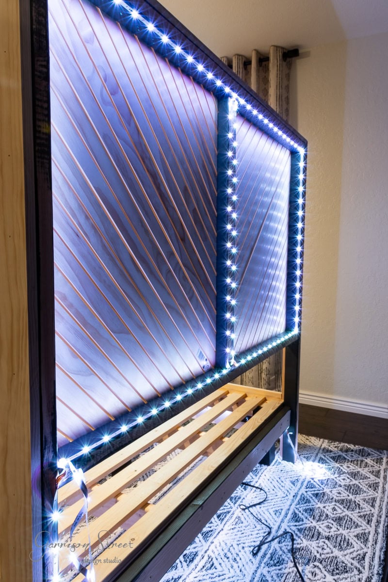 A wooden headboard with a slatted base is adorned with diagonal wires and bordered by bright LED lights. This DIY structure rests on a patterned rug and is set against a light-colored wall, perfect for adding a unique touch to your queen bed.