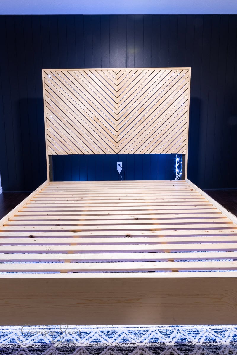 A wooden bed frame featuring a herringbone patterned headboard, ideal for a queen-sized DIY project, stands against a dark wall. Soft LED lights gently illuminate the base of the bed, which rests gracefully on a patterned rug.