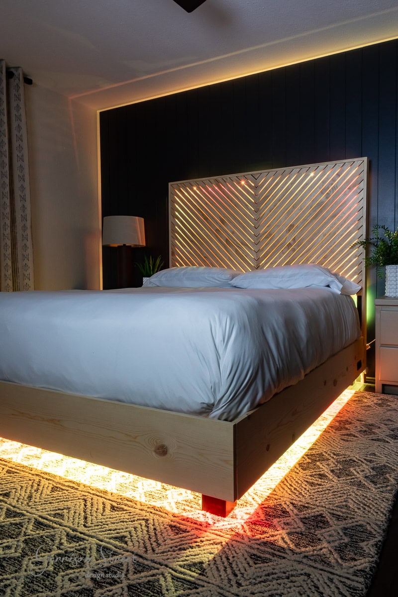 A modern bedroom showcases a DIY wooden bed with LED lighting gracing the headboard, perfect for a queen-sized mattress. The white bedding complements the patterned rug, while soft lighting creates a cozy atmosphere, accentuated by a plant and lamp in the background.