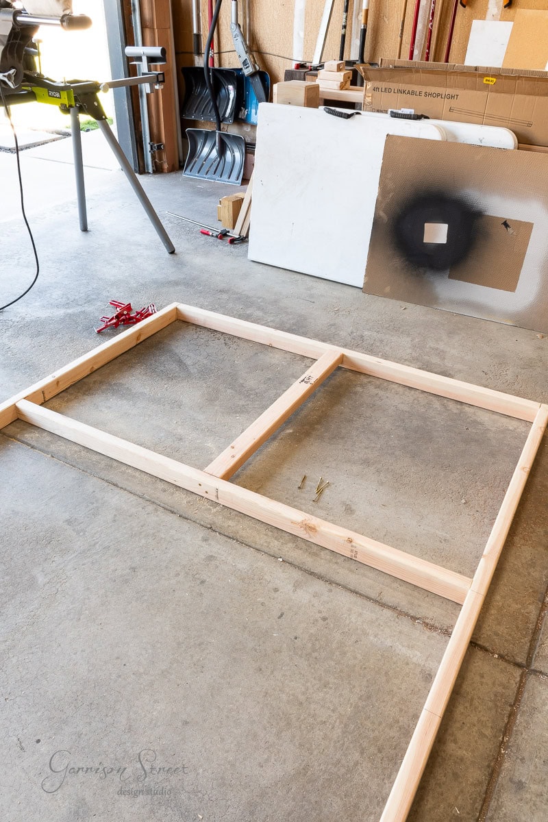 In a garage with a wooden frame on the concrete floor, tools and materials surround the scene, including a saw and a board with a black square. It's the perfect setup for your next DIY project, like crafting that headboard for your queen bed.