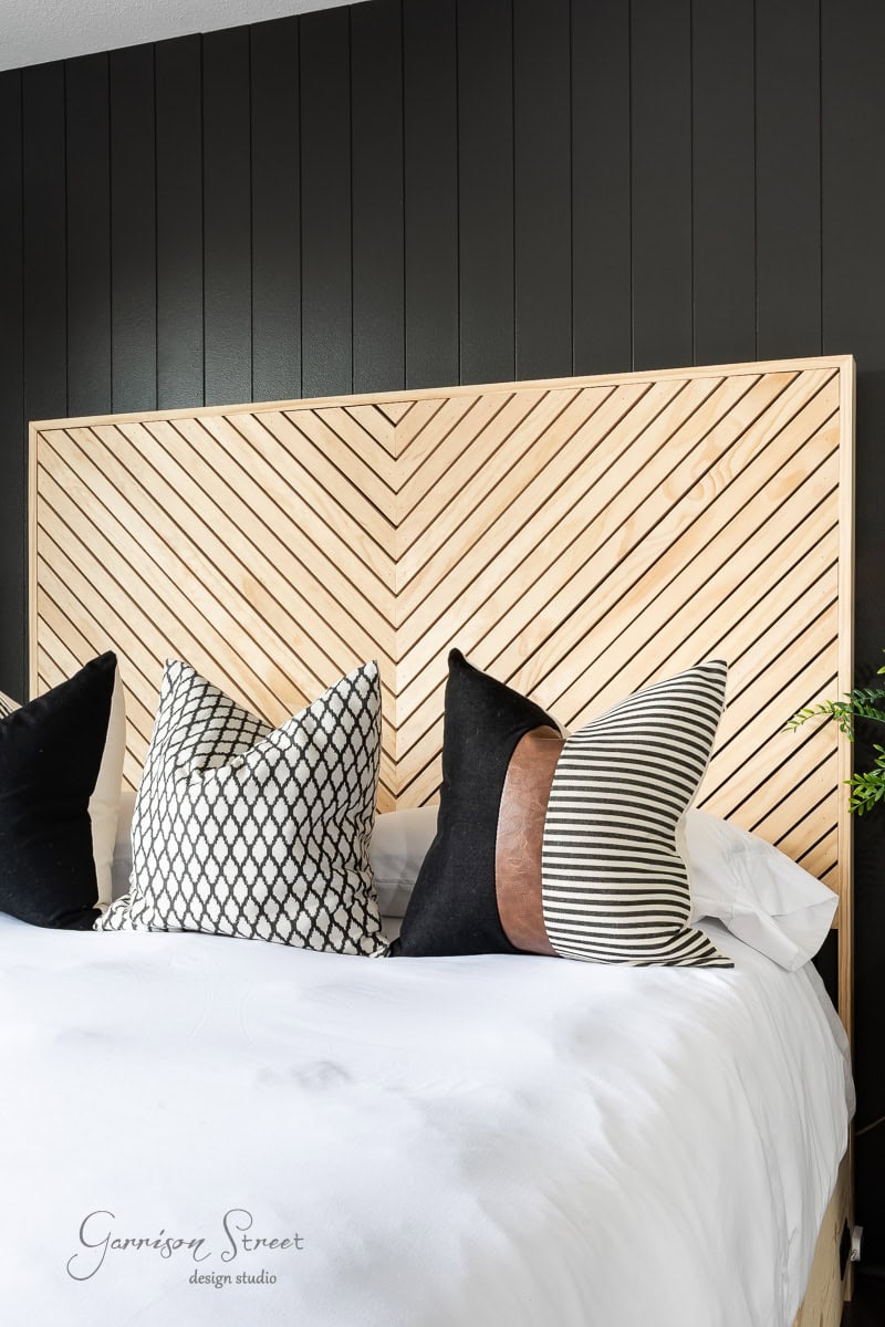 A modern bedroom showcases a DIY, chevron-patterned wood queen headboard against a dark paneled wall. The bed is adorned with a white sheet and several decorative pillows featuring black, white, and brown stripes along with geometric designs.