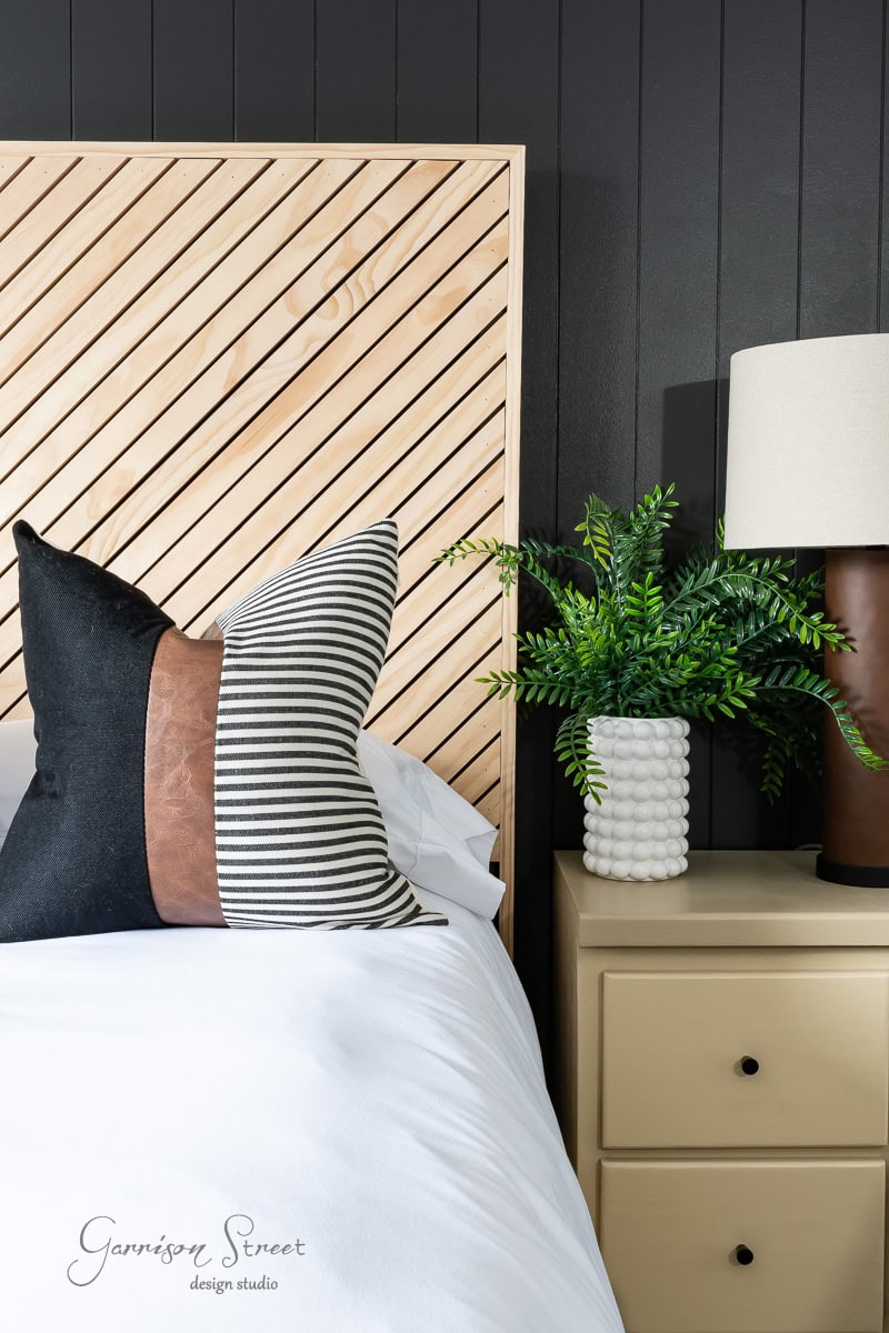 A modern bedroom showcases a DIY wooden diagonal slat headboard, queen-sized, against a dark wall. It includes striped and solid black pillows on a white bed, a beige nightstand with a white textured vase of green foliage, and a brown lamp with a cream shade.
