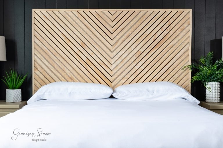 A modern queen bed with a white comforter and pillows is positioned against a striking wooden chevron-patterned headboard. Two bedside tables hold potted green plants and lamps, adding a fresh touch to the sleek bedroom decor, perfect for those considering a DIY approach.