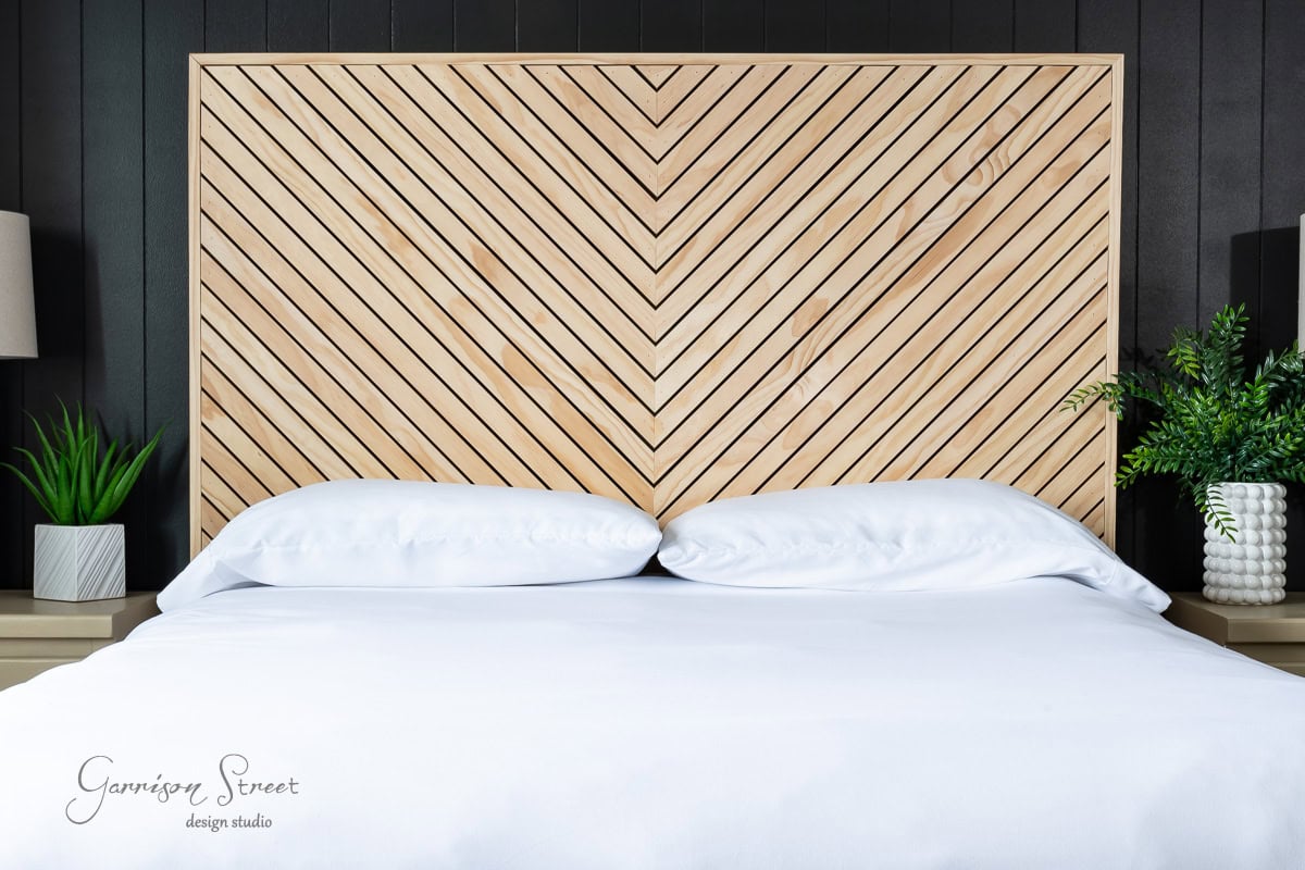 A modern queen bed with a white comforter and pillows is positioned against a striking wooden chevron-patterned headboard. Two bedside tables hold potted green plants and lamps, adding a fresh touch to the sleek bedroom decor, perfect for those considering a DIY approach.